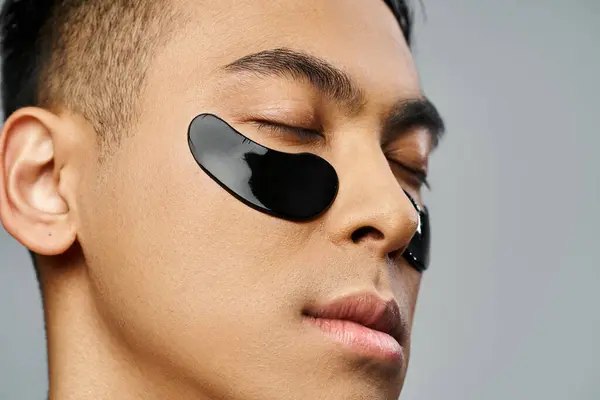 Hombre asiático guapo en una rutina de belleza, con un parche de ojo negro en un entorno de estudio gris. - foto de stock