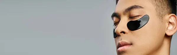 Bonito homem asiático ostentando um remendo olho preto em um estúdio cinza durante uma rotina de beleza e cuidados com a pele. — Fotografia de Stock