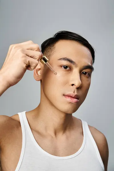 Un hombre asiático guapo usando suero durante una rutina de belleza en un estudio gris. - foto de stock