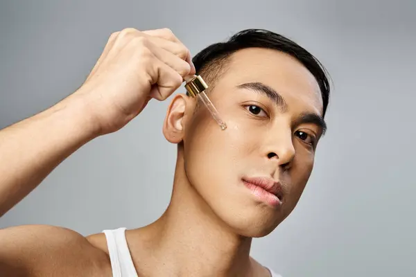 Um homem asiático bonito segurando pipeta com soro perto da bochecha durante uma rotina de beleza e cuidados com a pele em um estúdio cinza. — Fotografia de Stock