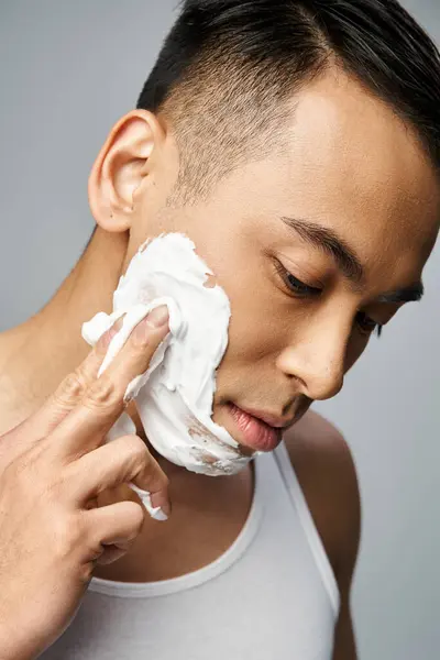 Красивий азіатський чоловік наносить піну для гоління на обличчя в сірій кімнаті. — стокове фото