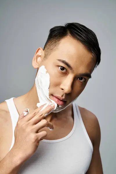 A handsome Asian man with shaving foam on his face in a grey studio, carefully shaving — Stock Photo