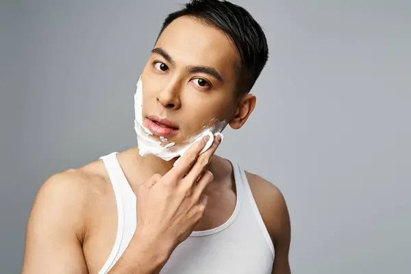 Beau asiatique avec mousse à raser sur le visage, se rasant méticuleusement avec un rasoir dans un studio gris. — Photo de stock