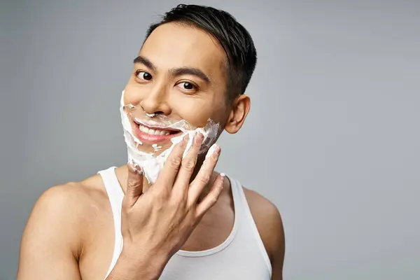 Красивий азіатський чоловік з піною для гоління на обличчі, ретельно голиться бритвою в сірій студії. — стокове фото