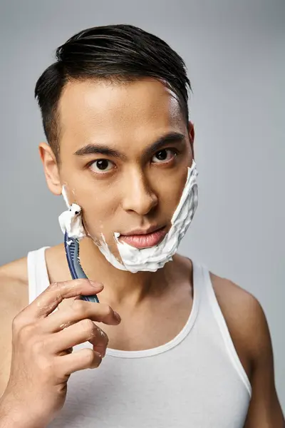 Un homme asiatique avec de la mousse à raser sur le visage se rase soigneusement avec un rasoir dans un cadre de studio gris serein. — Photo de stock