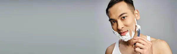 Un uomo asiatico con schiuma da barba sul viso radendosi accuratamente i peli del viso con un rasoio in uno studio grigio. — Foto stock