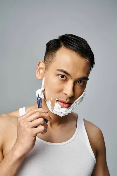 A stylish Asian man in black attire gracefully shaves his face with a razor in a vibrant red studio setting. — Stock Photo