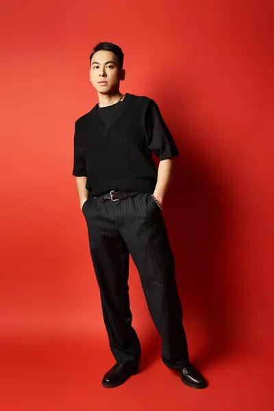 Un bel homme asiatique élégant se tient en confiance en tenue noire sur un fond rouge audacieux dans un cadre de studio. — Photo de stock