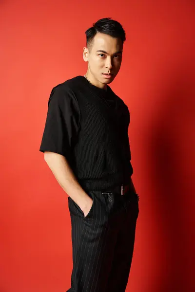 Um homem asiático elegante em trajes pretos confiantemente fica na frente de uma parede vermelha vibrante em um estúdio. — Fotografia de Stock