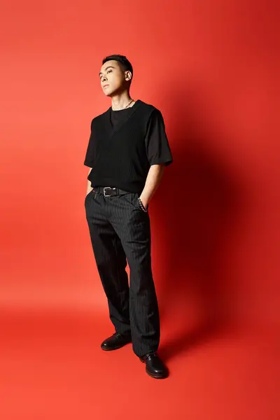 Модный азиат в черном уверенно стоит перед яркой красной стеной в студии. — стоковое фото