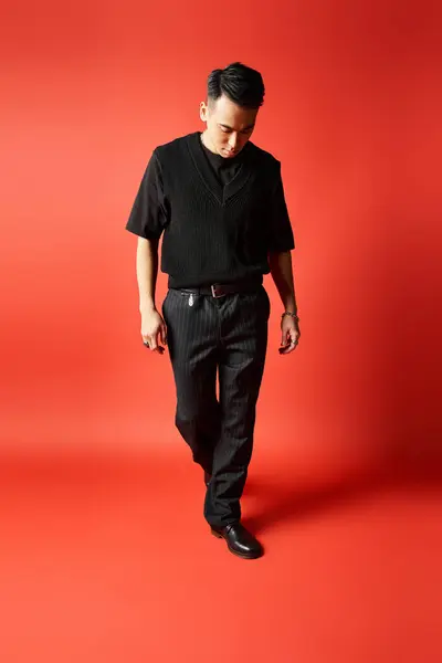 A stylish, handsome Asian man in black attire stands confidently in front of a vibrant red background in a studio. — Stock Photo