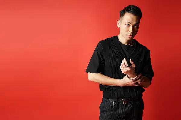 Um homem asiático elegante e bonito vestido com trajes pretos confiantemente fica na frente de uma impressionante parede vermelha em um estúdio. — Fotografia de Stock