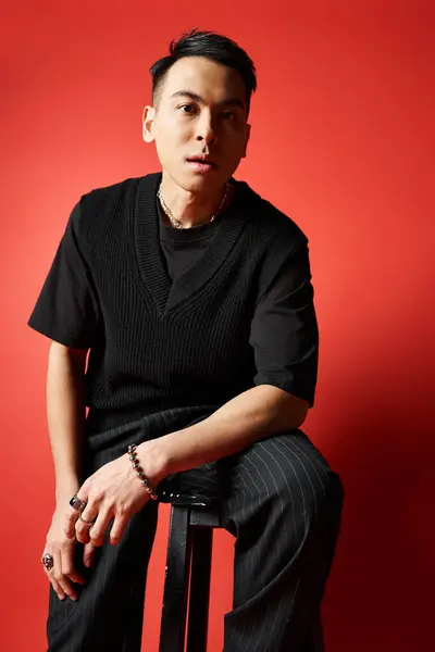 Un uomo asiatico elegante in abito nero si siede su uno sgabello di fronte a un muro rosso sorprendente in un ambiente di studio. — Foto stock
