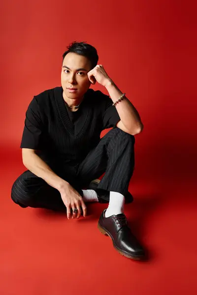 Handsome Asian man in black attire sitting on the ground, wearing black shoes, against a bold red studio background. — Stock Photo