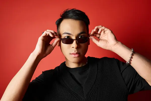 A stylish Asian man in black shirt and sunglasses poses confidently on a vibrant red background. — Stock Photo