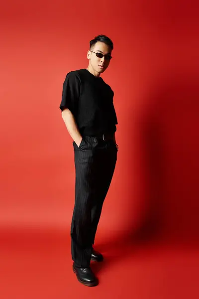 A handsome, stylish Asian man wearing a black shirt and black pants in a studio setting, standing confidently against a red background. — Stock Photo