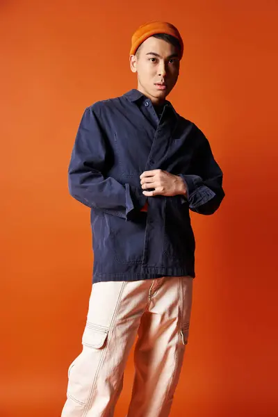 Ein gut aussehender asiatischer Mann in stylischer Kleidung steht selbstbewusst vor auffallend orangefarbenem Hintergrund in einem Studio-Setting. — Stockfoto