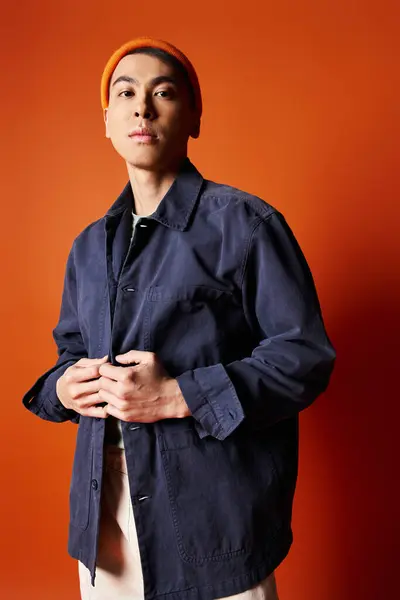 A handsome Asian man stands confidently in front of a vibrant orange wall in a studio setting, showcasing his stylish attire. — Stock Photo