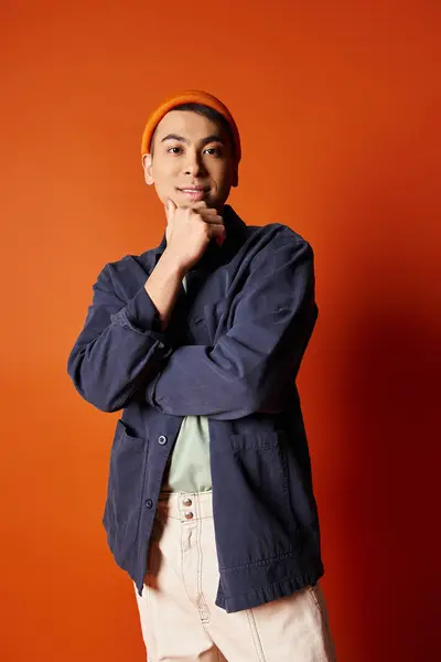 Un bell'uomo asiatico si trova fiducioso di fronte a una parete arancione brillante in un abbigliamento elegante. — Foto stock