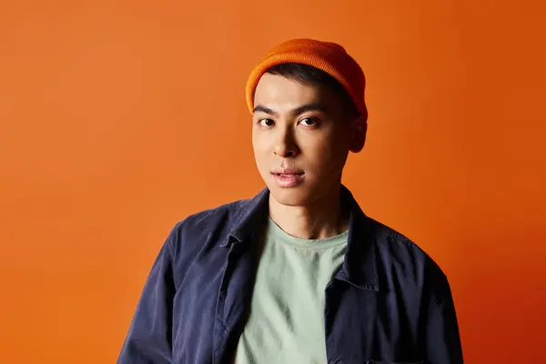 Ein gutaussehender asiatischer Mann in leuchtend blauer Jacke und orangefarbenem Hut steht selbstbewusst vor orangefarbenem Hintergrund in einem Studio. — Stockfoto