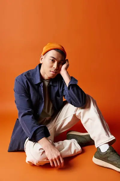 A handsome Asian man in stylish attire sits on the ground wearing a hat against an orange studio backdrop. — Stock Photo