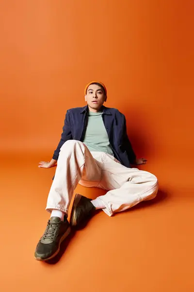 Un hombre asiático elegante, guapo, se sienta con las piernas cruzadas sobre un fondo naranja. - foto de stock