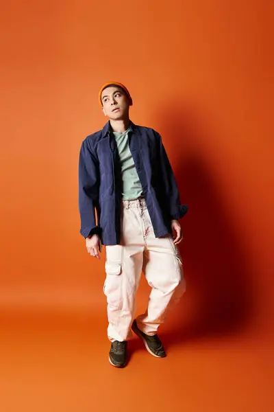 A handsome Asian man dressed in stylish attire stands confidently in front of a vivid orange wall in a studio setting. — Stock Photo