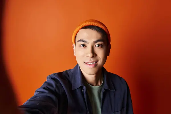Handsome Asian man dressed in stylish attire taking a selfie while wearing an orange hat in a vibrant studio setting. — Stock Photo