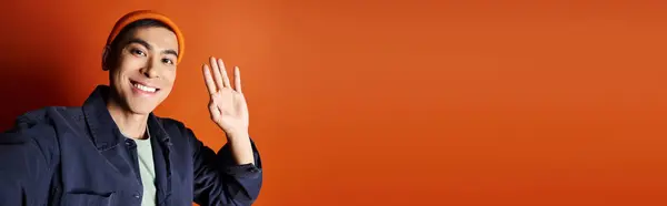 A stylish Asian man in a blue jacket is gesturing with his hand against a vibrant orange background in a studio setting. — Stock Photo