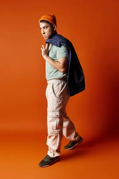 A handsome Asian man in stylish attire carries a backpack on his back against an orange background in a studio setting. — Stock Photo