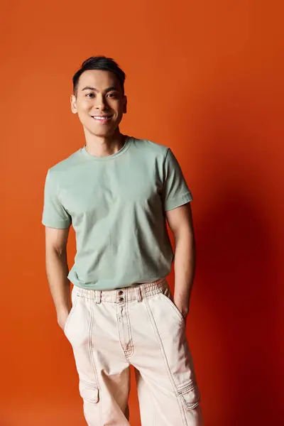 A fashionable Asian man in stylish attire standing confidently in front of a vibrant orange wall in a studio setting. — Stockfoto