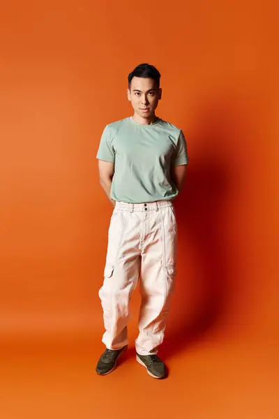 Красивый азиатский мужчина в стильной одежде уверенно стоит перед ярким оранжевым фоном в студии. — стоковое фото
