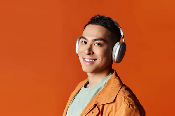 Un homme asiatique élégant en tenue élégante sourit tout en portant des écouteurs dans un cadre studio sur un fond orange. — Photo de stock