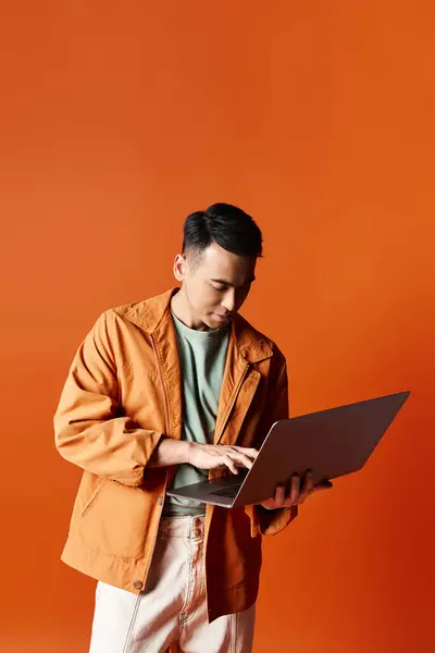 A stylish Asian man in an orange jacket engrossed in working on a laptop in a studio setting. — Stock Photo