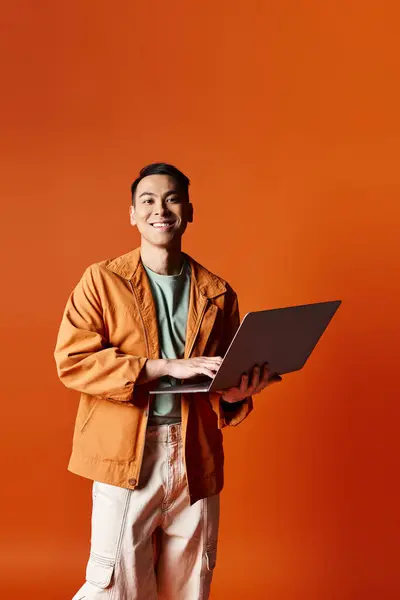 Un hombre asiático guapo, vestido con un atuendo elegante, sosteniendo un ordenador portátil mientras está de pie sobre un fondo naranja en un estudio. - foto de stock