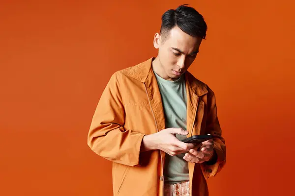 A handsome Asian man in a stylish orange jacket engrossed in his cell phone. — Stock Photo