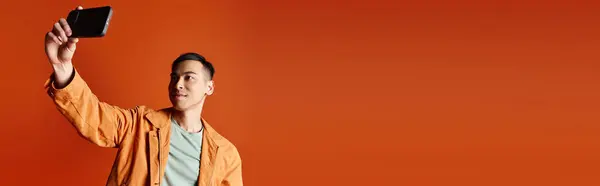 Un hombre asiático guapo con un atuendo elegante sosteniendo un teléfono celular en el aire contra un fondo naranja. - foto de stock