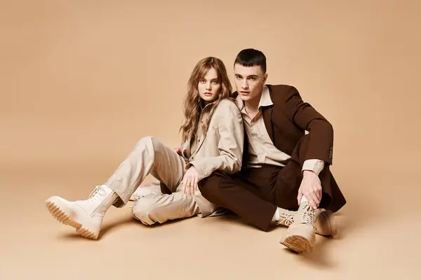 Atraente casal chique em ternos elegantes sentado no chão e olhando para a câmera em pano de fundo pastel — Fotografia de Stock