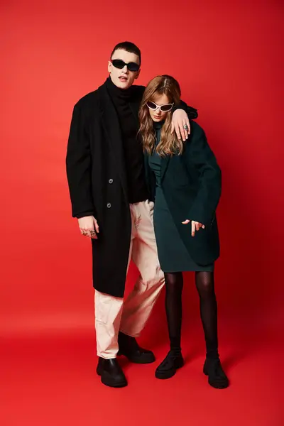 Beau couple à la mode en manteaux élégants avec des lunettes de soleil posant ensemble sur fond rouge — Photo de stock