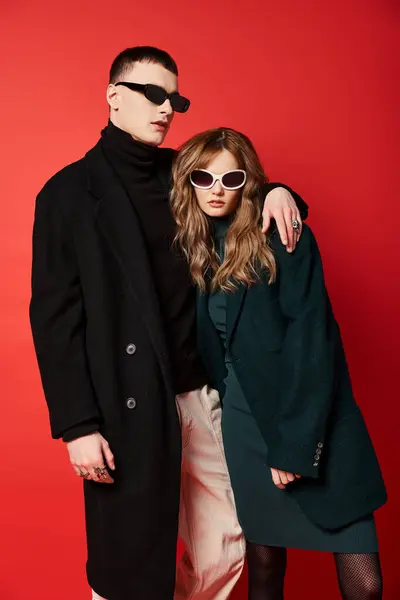 Pareja joven de moda en abrigos elegantes con gafas de sol de moda posando juntos sobre el telón de fondo rojo - foto de stock