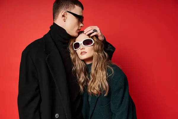 Fashionable young couple in stylish coats with trendy sunglasses posing together on red backdrop — Stock Photo