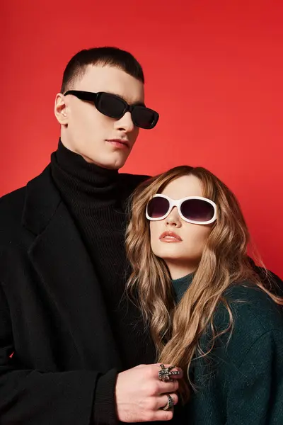 Модна молода пара в стильних пальто з модними сонцезахисними окулярами позує разом на червоному тлі — стокове фото