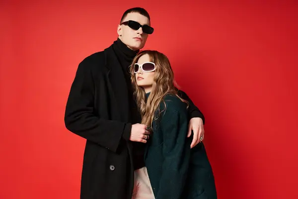 Sofisticato giovane coppia in eleganti cappotti con occhiali da sole in posa insieme su sfondo rosso — Foto stock