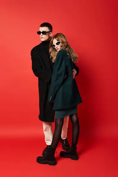 Sophisticated young couple in stylish coats with sunglasses posing together on red backdrop — Stock Photo