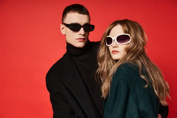 Витончена молода пара в стильних пальто з сонцезахисними окулярами позує разом на червоному тлі — стокове фото