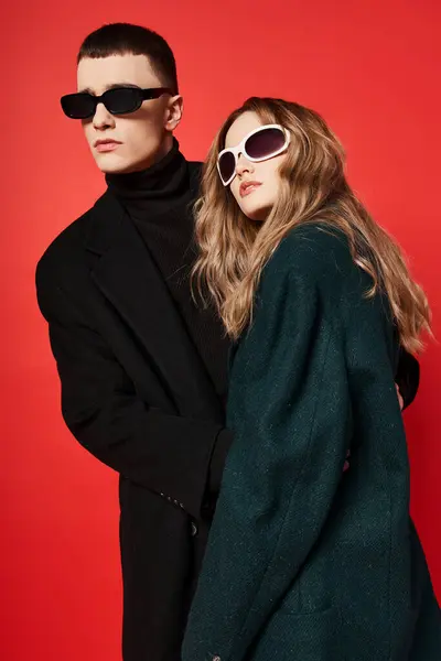 Bien vestida joven pareja en elegantes abrigos con gafas de sol posando juntos sobre fondo rojo - foto de stock
