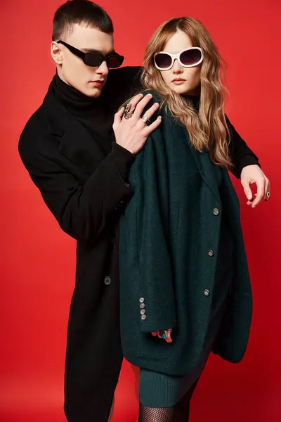 Bien vestida joven pareja en elegantes abrigos con gafas de sol posando juntos sobre fondo rojo - foto de stock