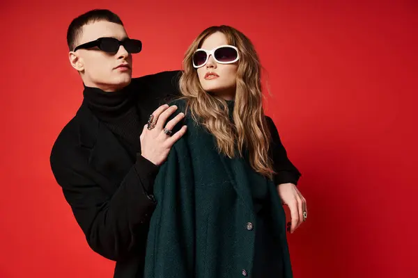 Elegante pareja joven en elegantes abrigos con gafas de sol de moda posando juntos sobre fondo rojo - foto de stock