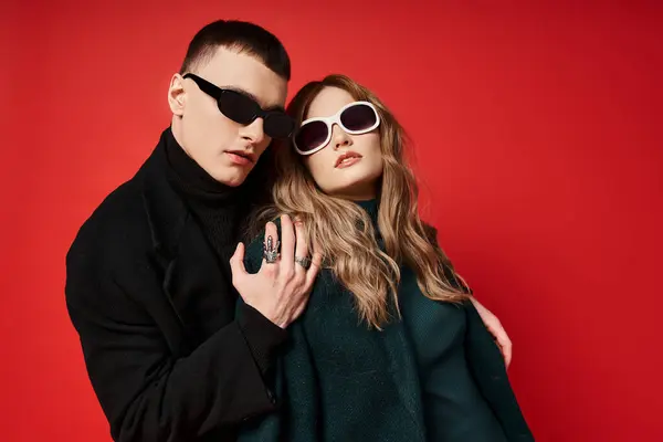 Élégant jeune couple en manteaux élégants avec des lunettes de soleil à la mode posant ensemble sur fond rouge — Photo de stock
