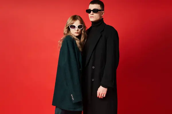 Elegante pareja joven en elegantes abrigos con gafas de sol de moda posando juntos sobre fondo rojo - foto de stock